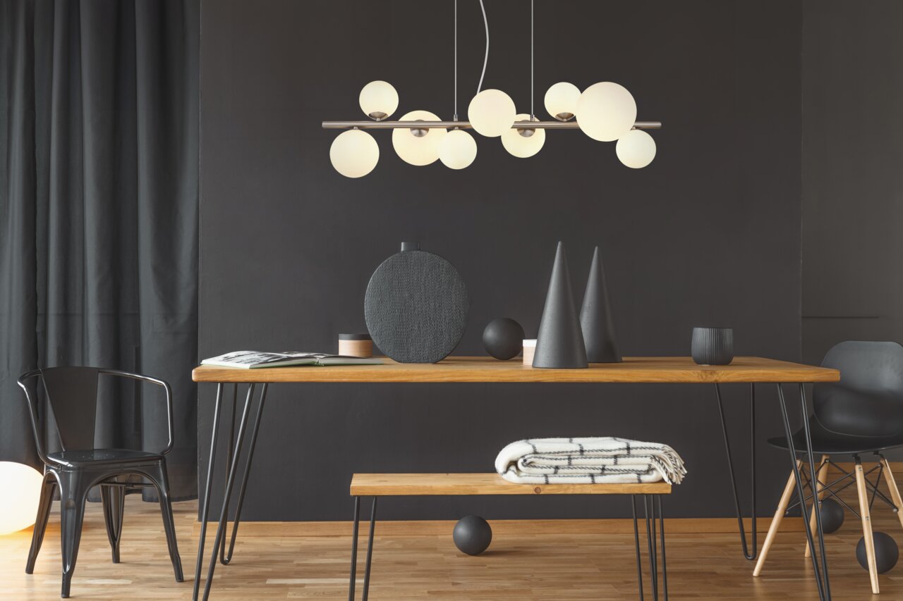 Moderne Hängeleuchte mit mehreren runden, weißen Glaskugeln, die von einer Metallstange hängen, in einem stilvoll eingerichteten Raum mit Holztisch und schwarzen Dekorationselementen. Perspektive: Frontalansicht.