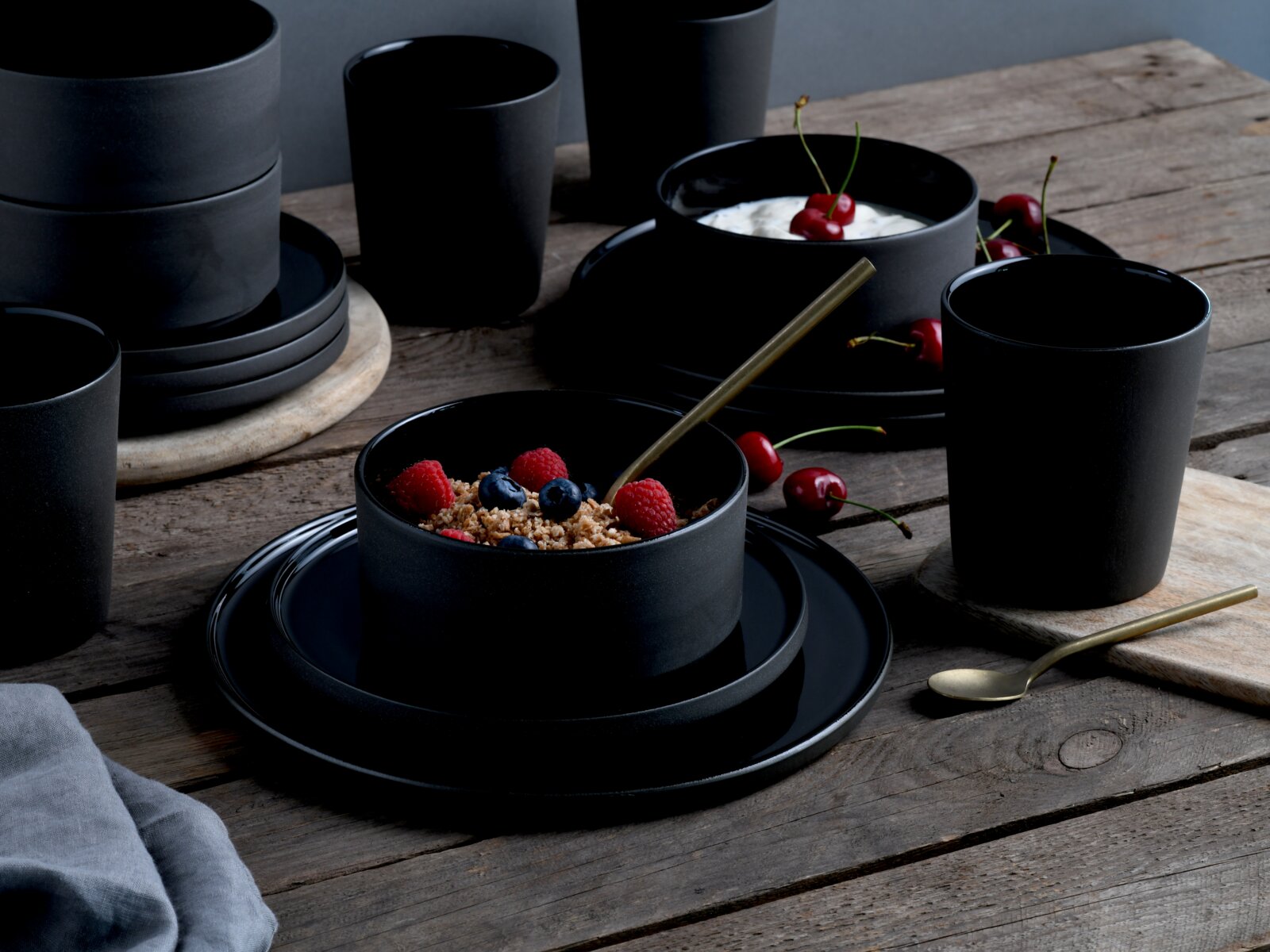 Creatable Kombiservice 8-tlg FUTURO Schwarzes, mattes Geschirrset mit Schalen, Tellern und Bechern auf rustikalem Holztisch, teilweise mit Müsli und Joghurt mit Kirschen angerichtet, aufgenommen in leicht schräger Seitenansicht von vorne.