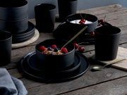 Schwarzes, mattes Geschirrset mit Schalen, Tellern und Bechern auf rustikalem Holztisch, teilweise mit Müsli und Joghurt mit Kirschen angerichtet, aufgenommen in leicht schräger Seitenansicht von vorne.
