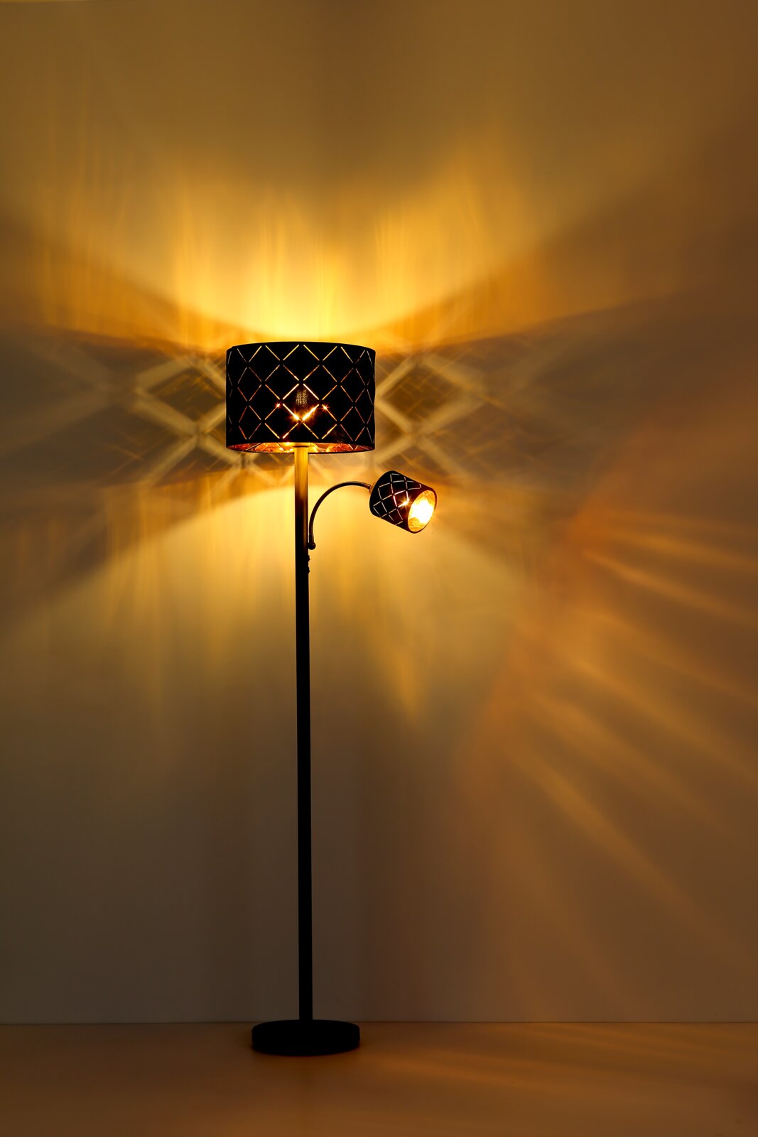 Stehleuchte mit zwei Leuchtkörpern, die warmes Licht und ein dekoratives Muster an die Wand projizieren, seitliche Perspektive.