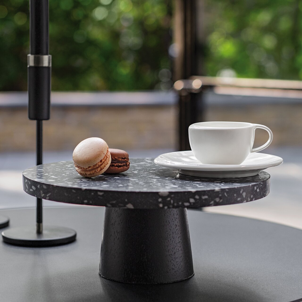 Weiße Kaffeeuntertasse NewMoon mit Tasse, seitliche Perspektive, auf einem dunklen Tisch mit zwei Macarons.