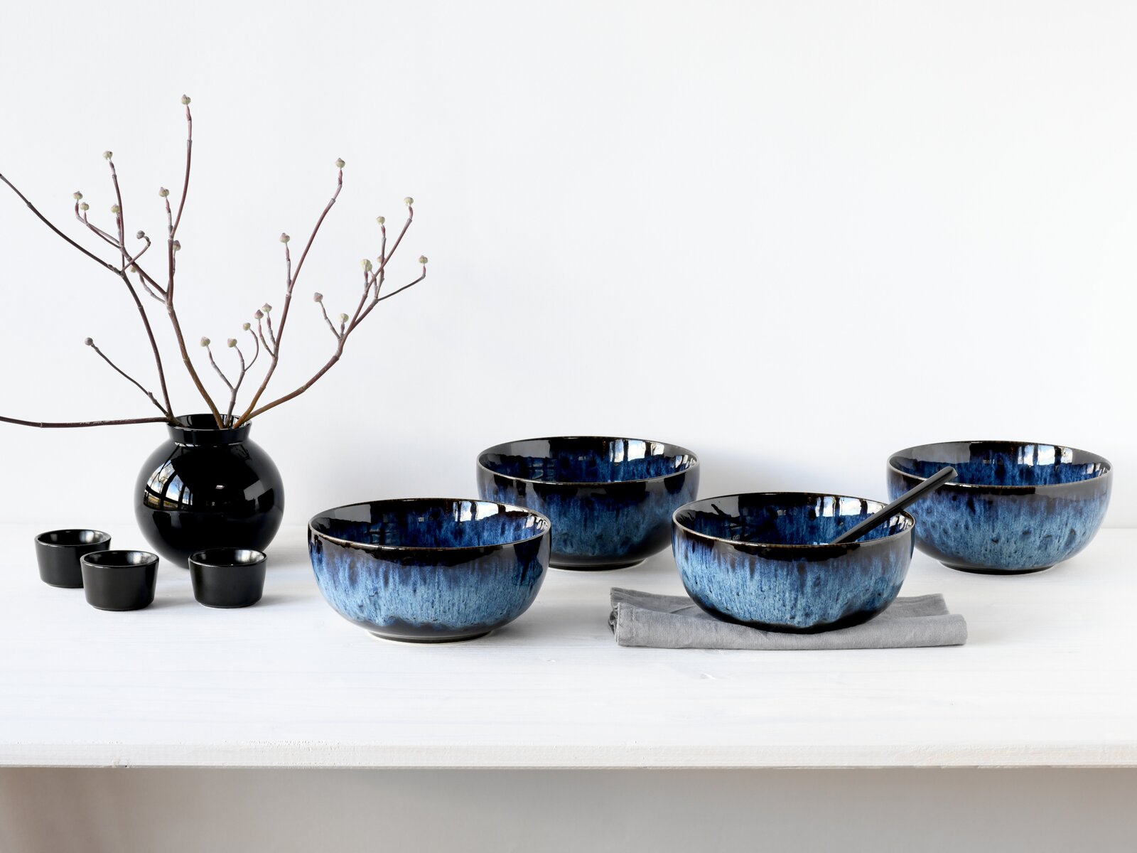 Blau-schwarzes Keramikschalen-Set in unterschiedlichen Größen auf weißem Tisch, eine Schale mit schwarzem Löffel auf grauem Tuch seitliche Frontansicht.