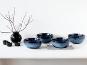 Blau-schwarzes Keramikschalen-Set in unterschiedlichen Größen auf weißem Tisch, eine Schale mit schwarzem Löffel auf grauem Tuch; seitliche Frontansicht.
