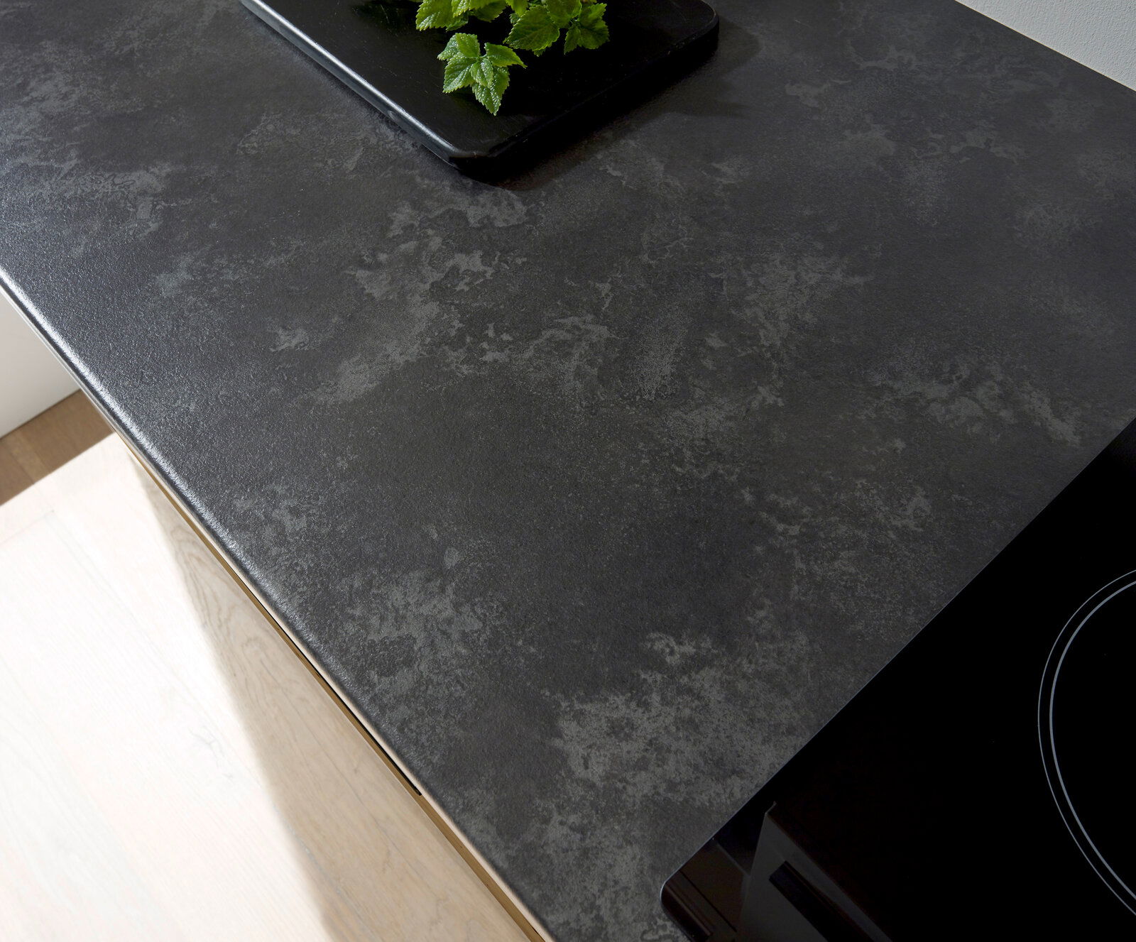 Draufsicht auf eine moderne, dunkle Küchenarbeitsplatte mit einem Kochfeld und einem Tablett mit grünen Kräutern.