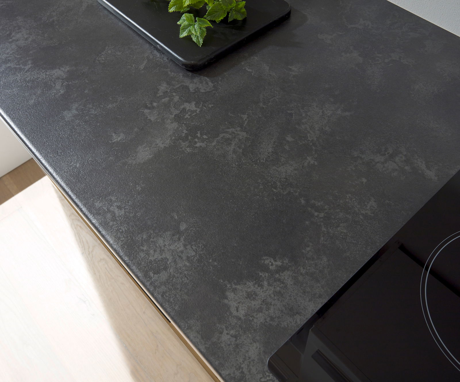 Draufsicht auf eine moderne, dunkle Küchenarbeitsplatte mit einem Kochfeld und einem Tablett mit grünen Kräutern.