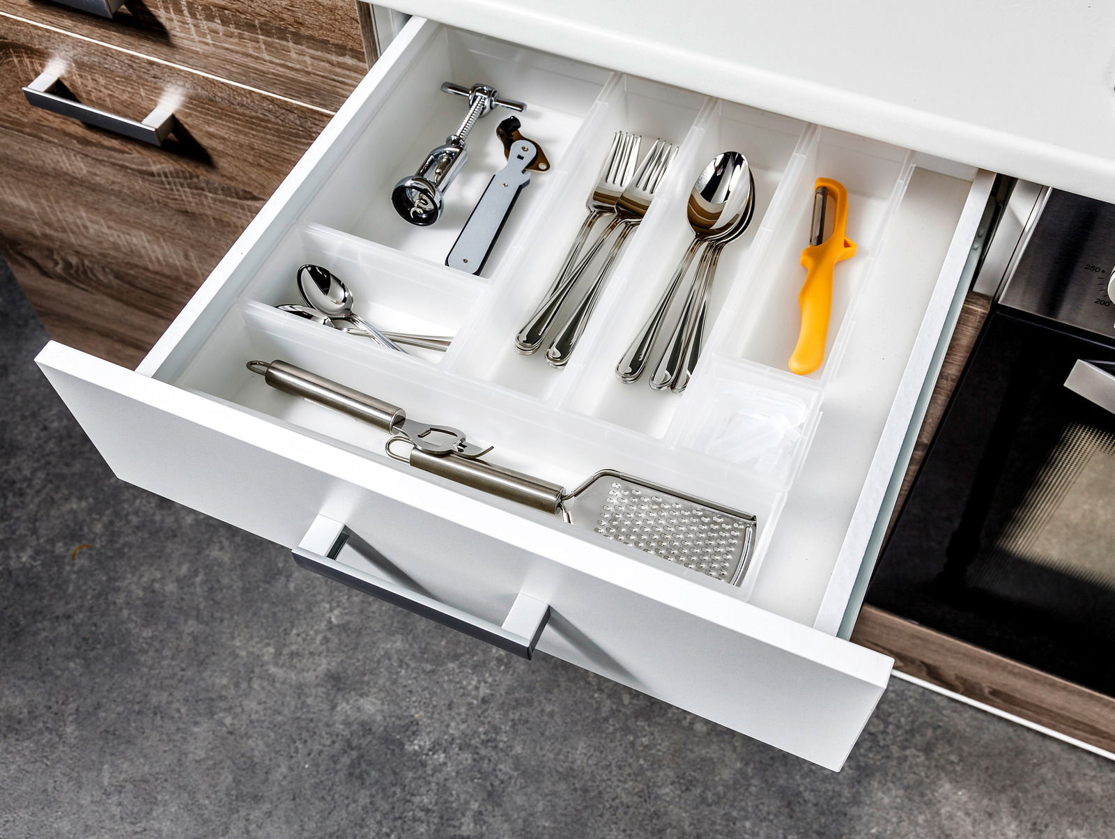 Offene Küchenschublade mit weißem Ordnungssystem, gefüllt mit Besteck und Küchenutensilien, von oben betrachtet.