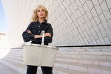 Frau mit einem Carrybag in Herringbone Sand vor einer modernen Architekturkulisse, Frontalansicht