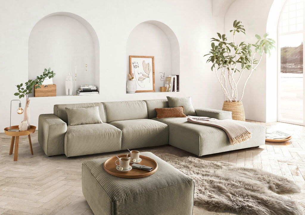 Modernes, helles Wohnzimmer mit einem großen, beigen Ecksofa in L-Form, dekoriert mit Kissen und einer Decke, umgeben von Pflanzen und stilvollen Accessoires. Perspektive von vorne links.