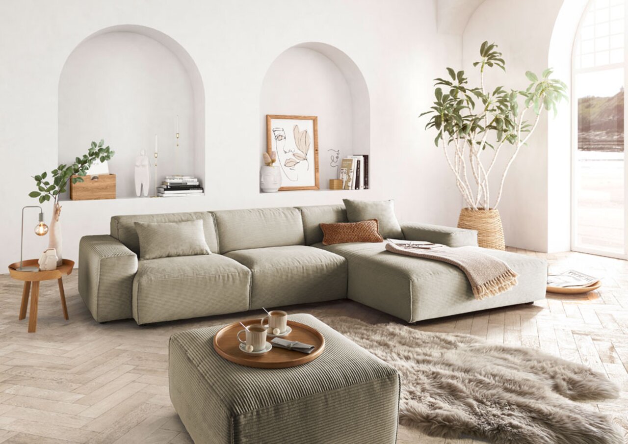 Modernes, helles Wohnzimmer mit einem großen, beigen Ecksofa in L-Form, dekoriert mit Kissen und einer Decke, umgeben von Pflanzen und stilvollen Accessoires. Perspektive von vorne links.