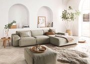 Modernes, helles Wohnzimmer mit einem großen, beigen Ecksofa in L-Form, dekoriert mit Kissen und einer Decke, umgeben von Pflanzen und stilvollen Accessoires. Perspektive von vorne links.