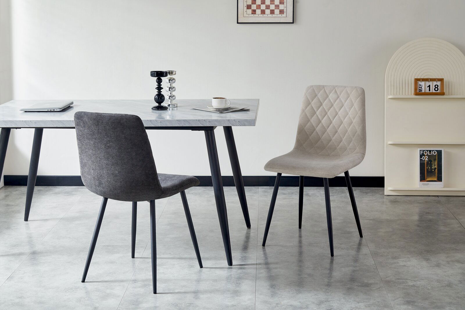 Zwei moderne Stühle mit schwarzen Beinen, einer in Grau und einer in Beige, stehen vor einem Tisch in einem minimalistischen Raum. Perspektive von vorne.