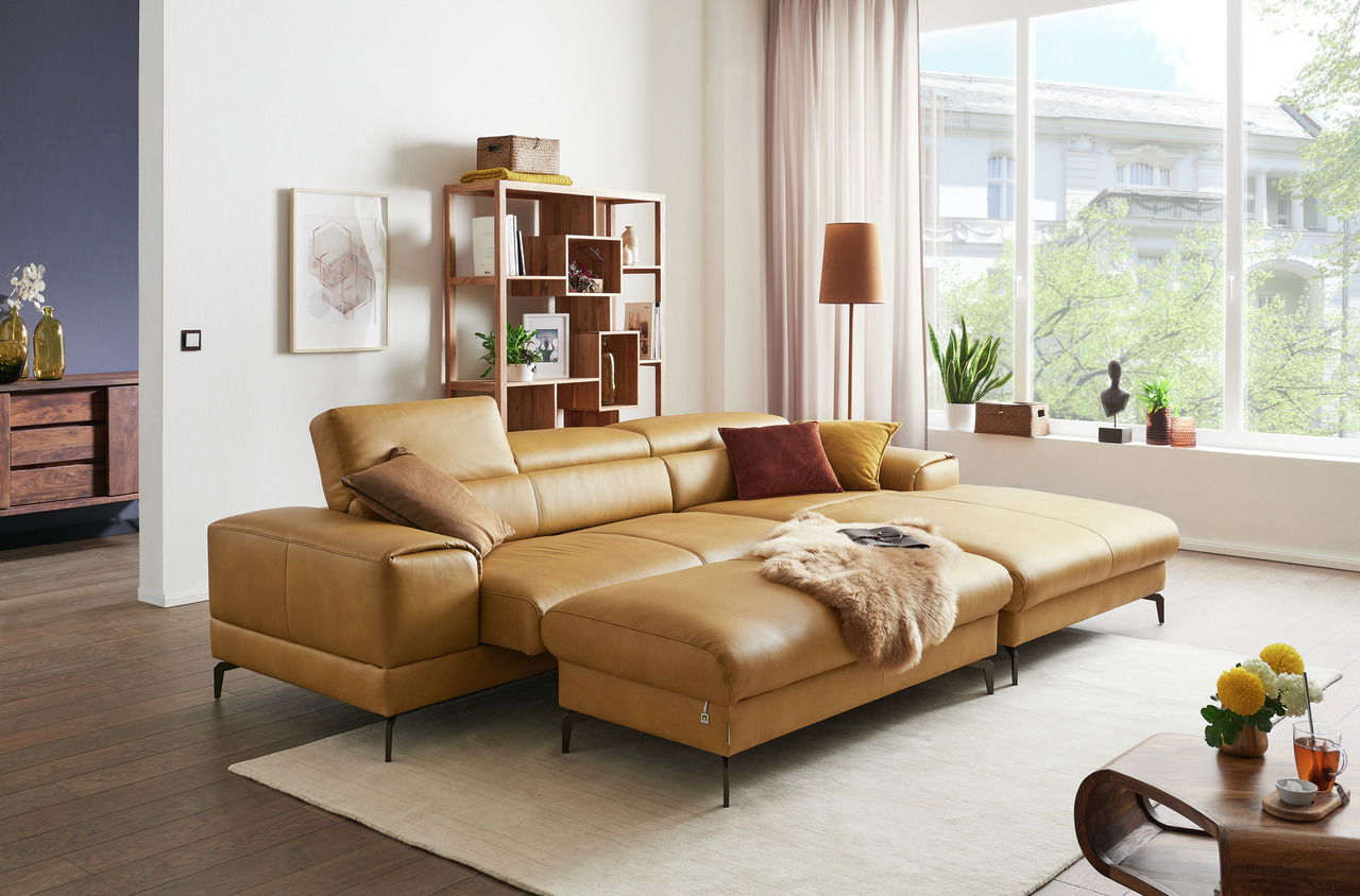 Ein modernes Wohnzimmer mit einem großen, senffarbenen Ledersofa in L-Form, das von vorne betrachtet wird. Das Sofa ist mit bunten Kissen und einer Felldecke dekoriert. Im Hintergrund sind ein Bücherregal, eine Stehlampe und große Fenster mit Blick auf gr