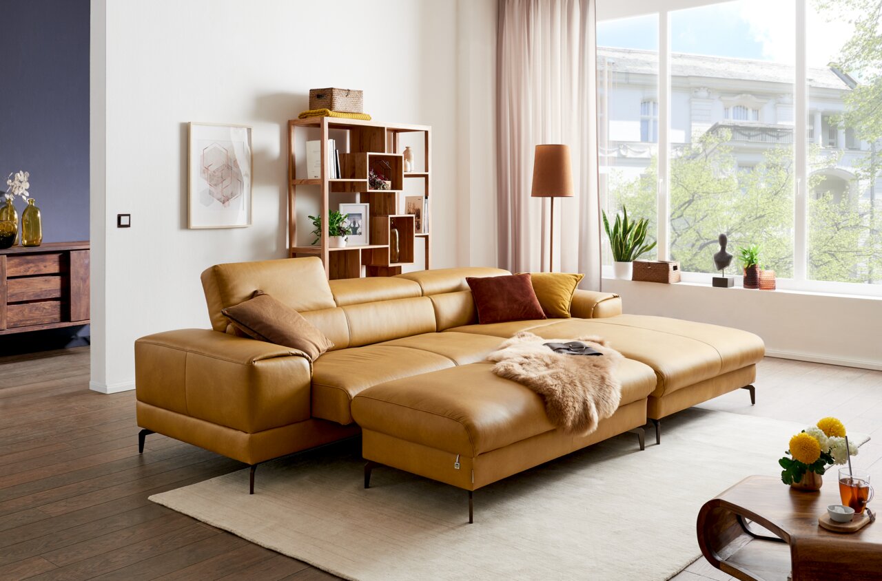 Ein modernes Wohnzimmer mit einem großen, senffarbenen Ledersofa in L-Form, das von vorne betrachtet wird. Das Sofa ist mit bunten Kissen und einer Felldecke dekoriert. Im Hintergrund sind ein Bücherregal, eine Stehlampe und große Fenster mit Blick auf gr