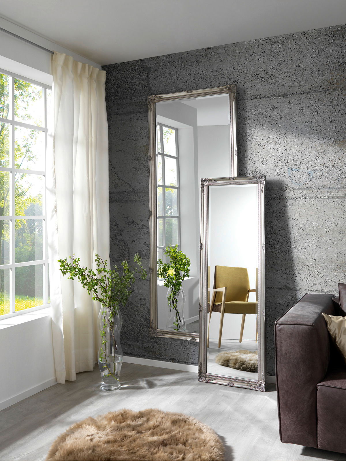 Zwei große, rechteckige Spiegel mit dekorativem Rahmen, die an einer grauen Wand in einem modernen Wohnzimmer lehnen. Der Raum ist hell und stilvoll eingerichtet mit einem großen Fenster, einem braunen Sofa und einem flauschigen Teppich. Die Spiegel refle