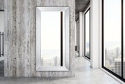 Großer rechteckiger Spiegel mit silbernem Rahmen, an einer modernen Betonwand montiert, mit Blick auf eine Stadtlandschaft durch große Fenster.