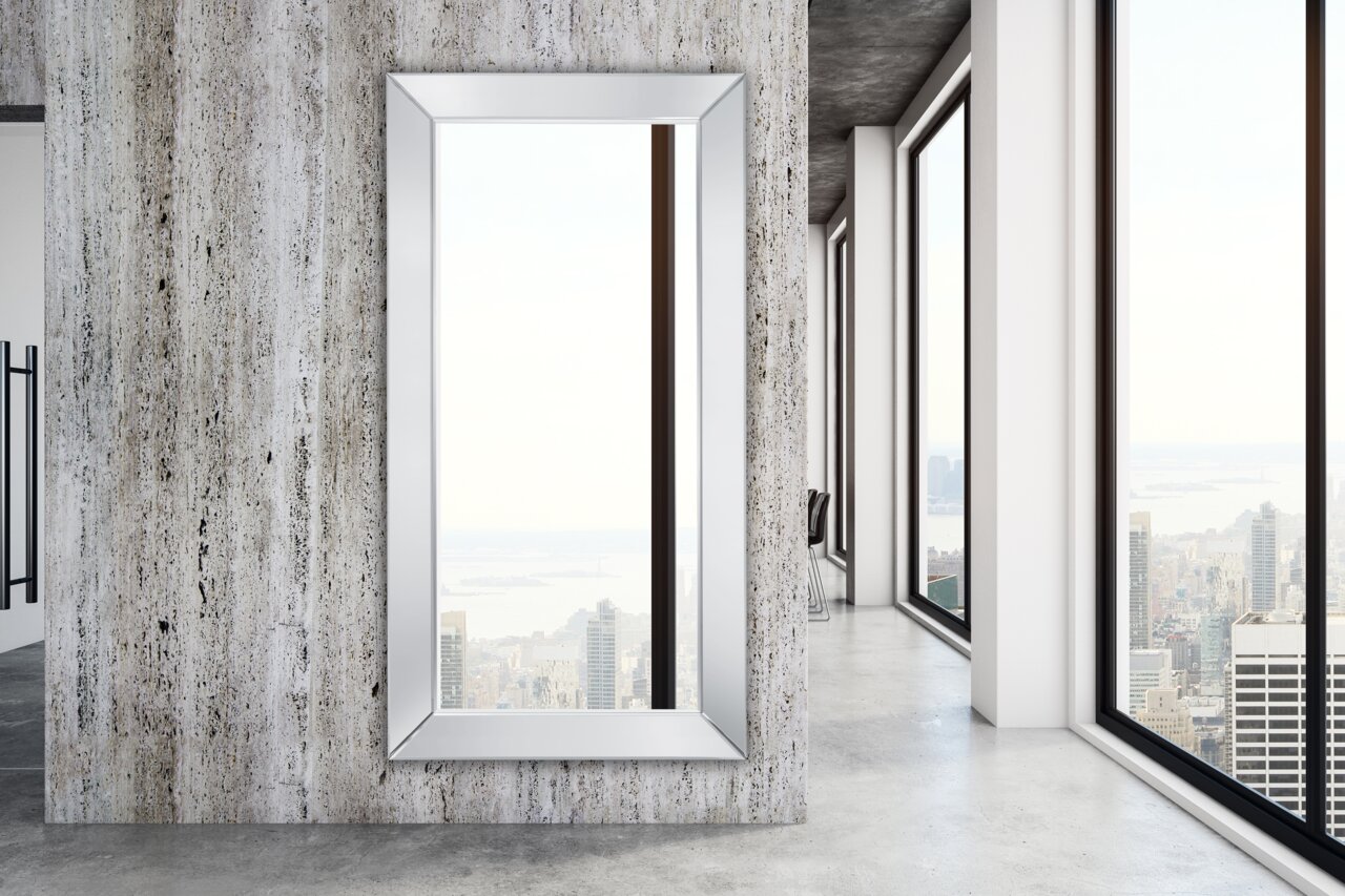 Großer rechteckiger Spiegel mit silbernem Rahmen, an einer modernen Betonwand montiert, mit Blick auf eine Stadtlandschaft durch große Fenster.