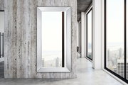 Großer rechteckiger Spiegel mit silbernem Rahmen, an einer modernen Betonwand montiert, mit Blick auf eine Stadtlandschaft durch große Fenster.