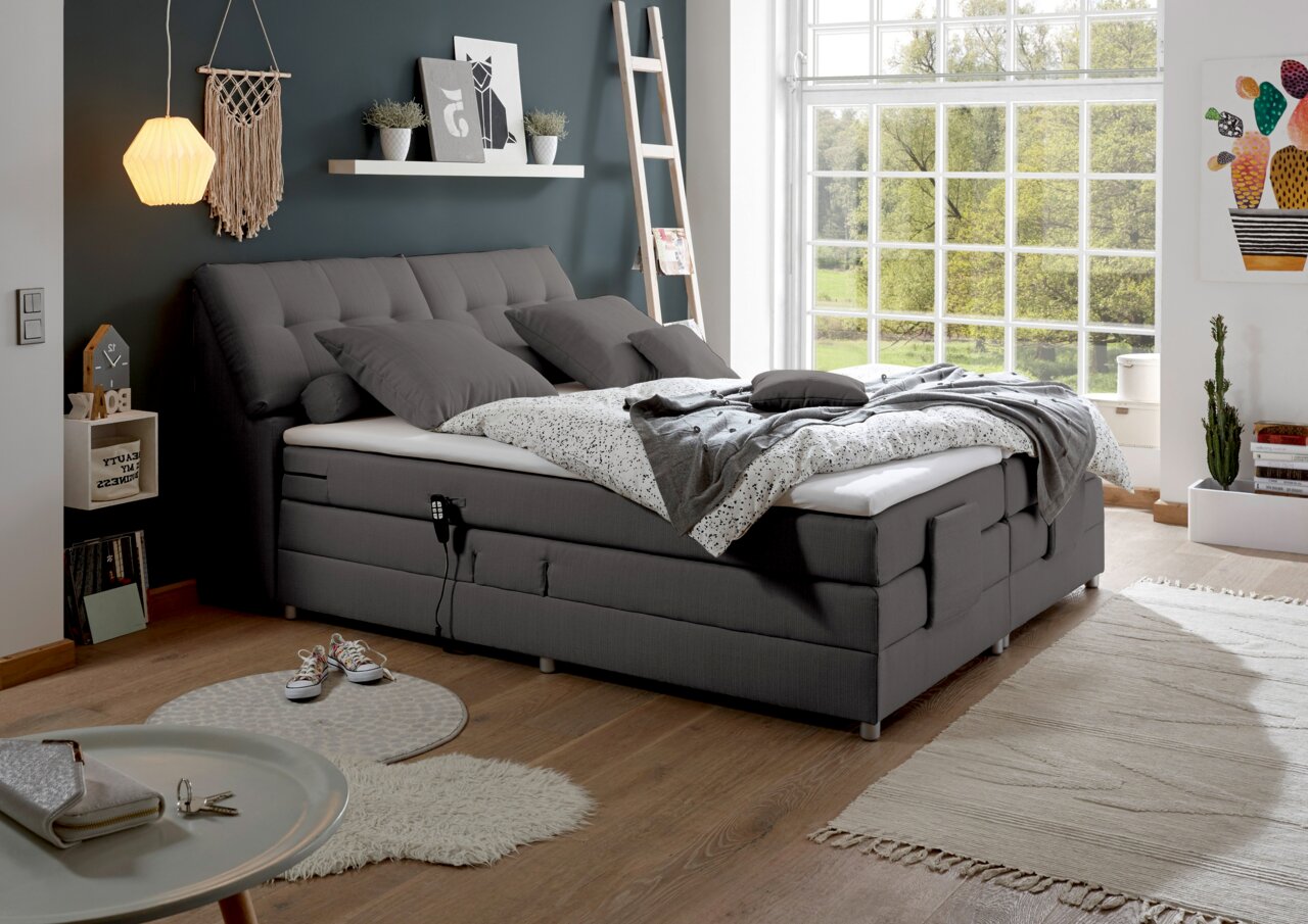 Gemütliches graues Boxspringbett in modernem Schlafzimmer mit Tageslicht, seitliche Perspektive