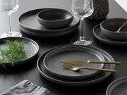 Elegantes Tellerset Uno Volkano 12-tlg. in dunklem Design, präsentiert auf einem Tisch mit goldfarbenem Besteck und Weingläsern, aus seitlicher Perspektive.