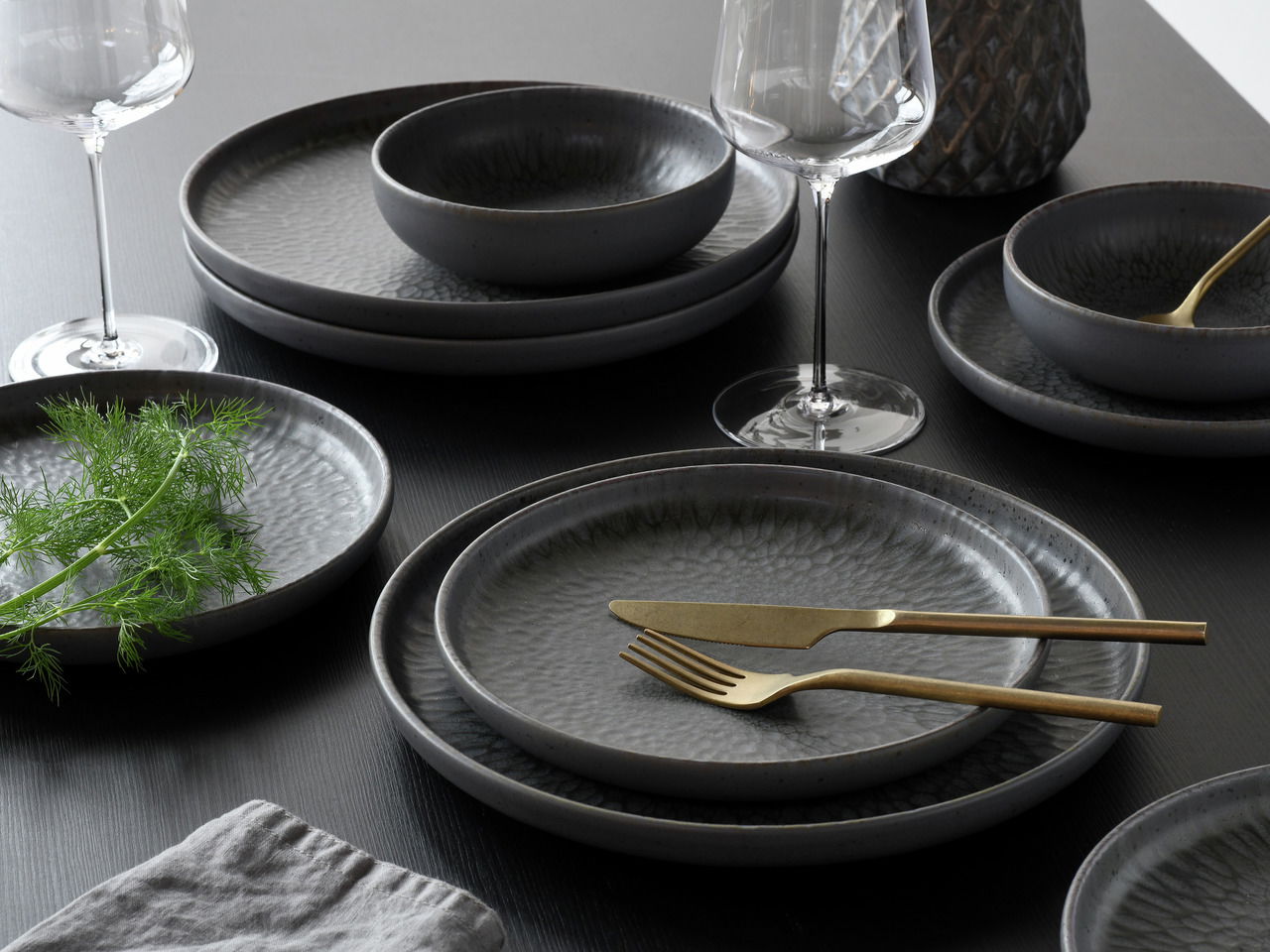 Elegantes Tellerset Uno Volkano 12-tlg. in dunklem Design, präsentiert auf einem Tisch mit goldfarbenem Besteck und Weingläsern, aus seitlicher Perspektive.
