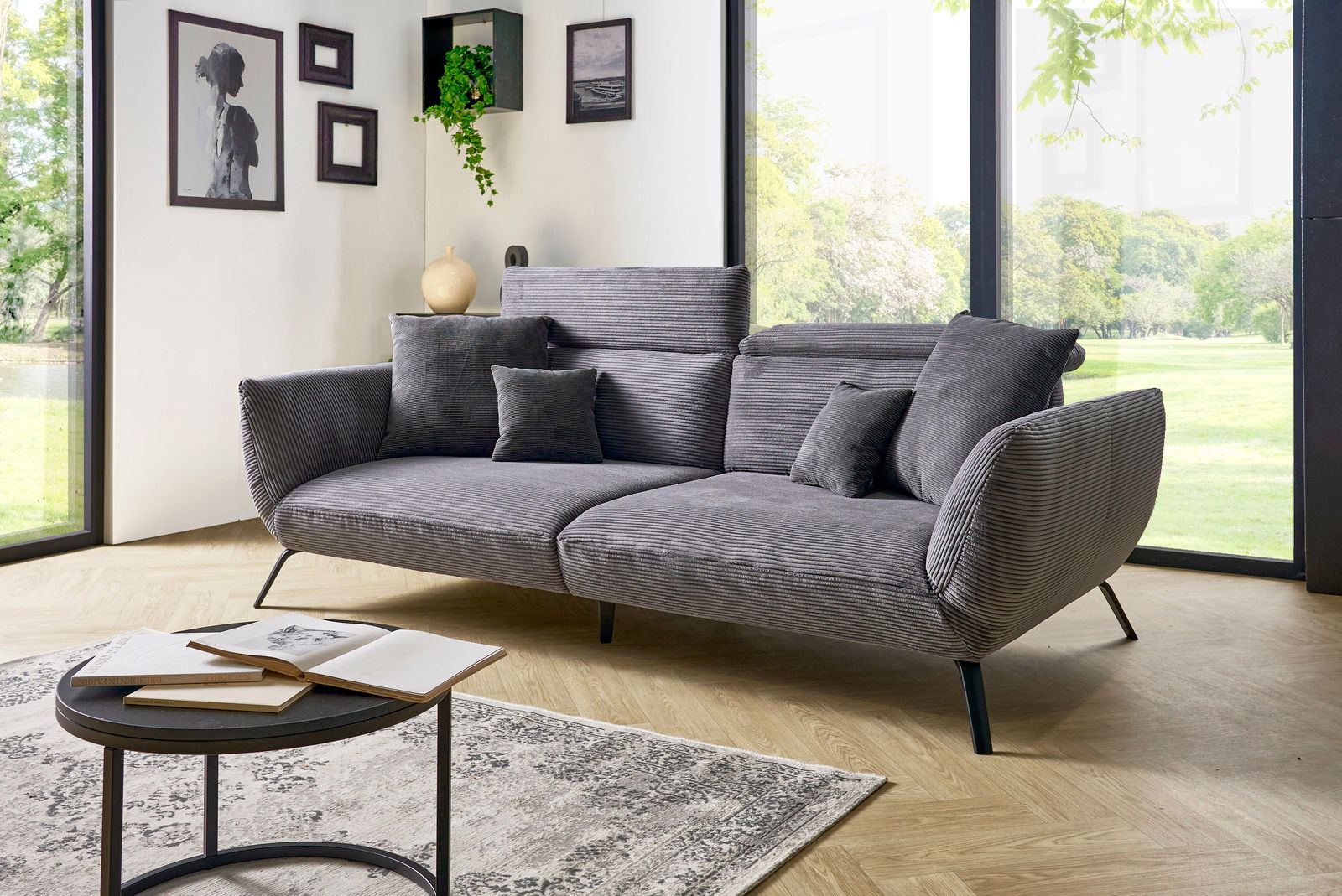 Graues Megasofa mit mehreren Kissen in einem modernen Wohnzimmer mit großen Fenstern und Blick ins Grüne, seitliche Perspektive.