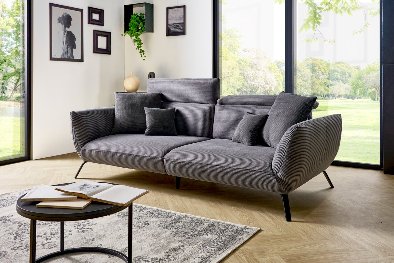 Graues Megasofa mit mehreren Kissen in einem modernen Wohnzimmer mit großen Fenstern und Blick ins Grüne, seitliche Perspektive.