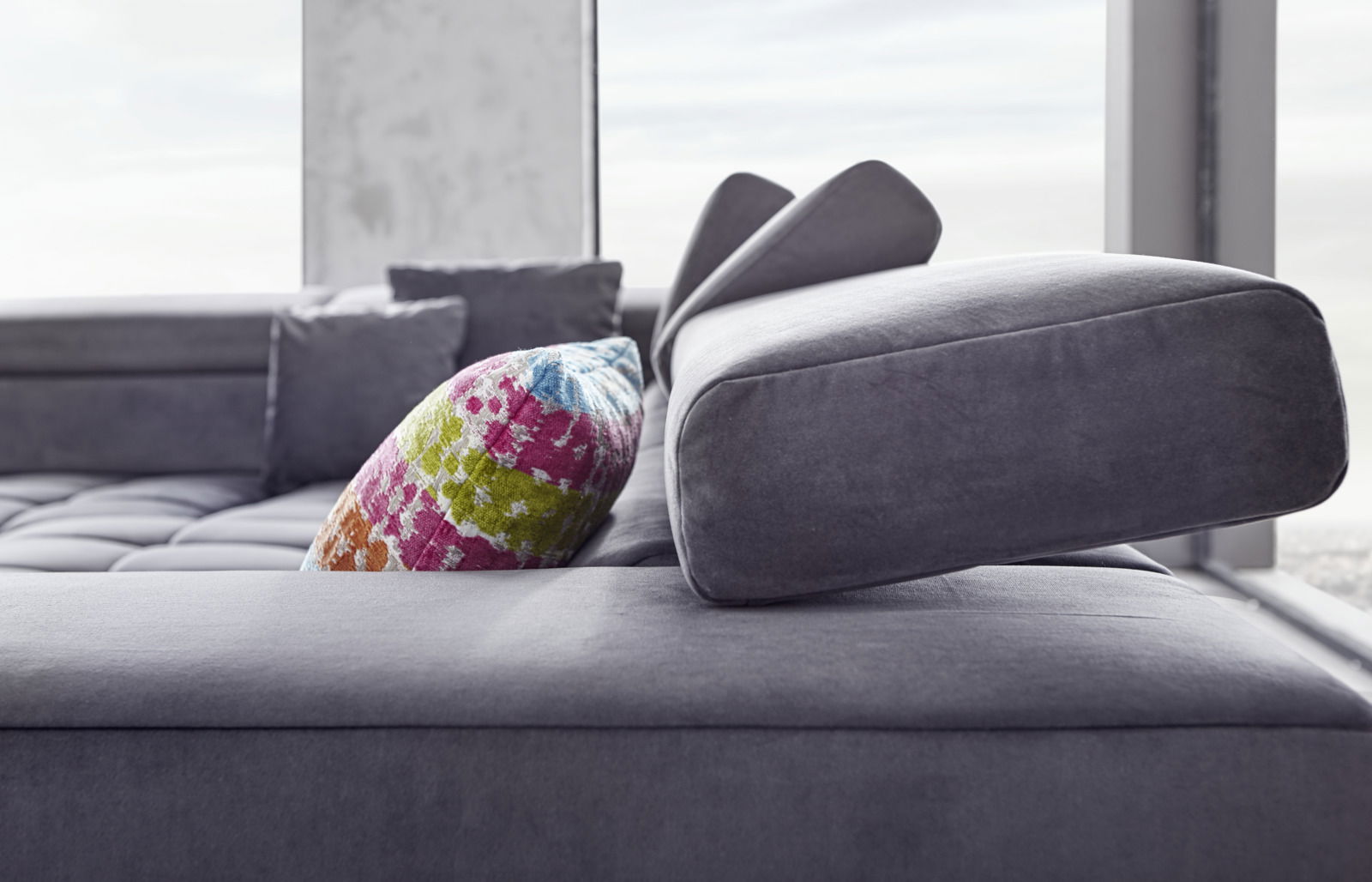 Nahaufnahme eines modernen Ecksofas in Grau mit verstellbarer Kopfstütze und einem bunten Kissen, seitliche Perspektive.