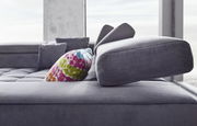 Nahaufnahme eines modernen Ecksofas in Grau mit verstellbarer Kopfstütze und einem bunten Kissen, seitliche Perspektive.