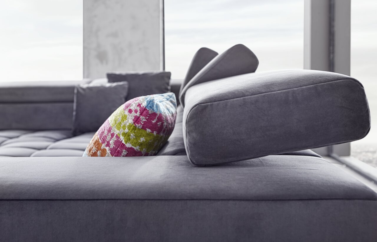 Nahaufnahme eines modernen Ecksofas in Grau mit verstellbarer Kopfstütze und einem bunten Kissen, seitliche Perspektive.
