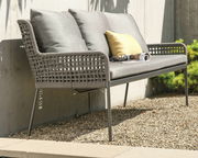 Seitliche Perspektive auf eine stilvolle Dining Bank mit grauem Geflecht und Kissen, platziert auf einer Terrasse mit Kiesboden.