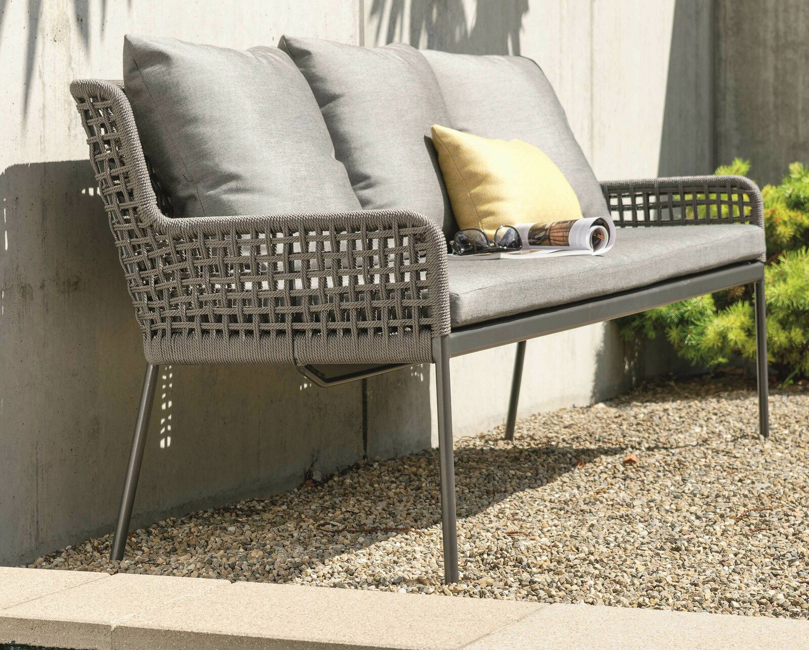 Seitliche Perspektive auf eine stilvolle Dining Bank mit grauem Geflecht und Kissen, platziert auf einer Terrasse mit Kiesboden.