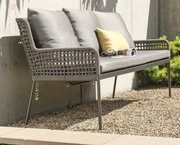 Seitliche Perspektive auf eine stilvolle Dining Bank mit grauem Geflecht und Kissen, platziert auf einer Terrasse mit Kiesboden.