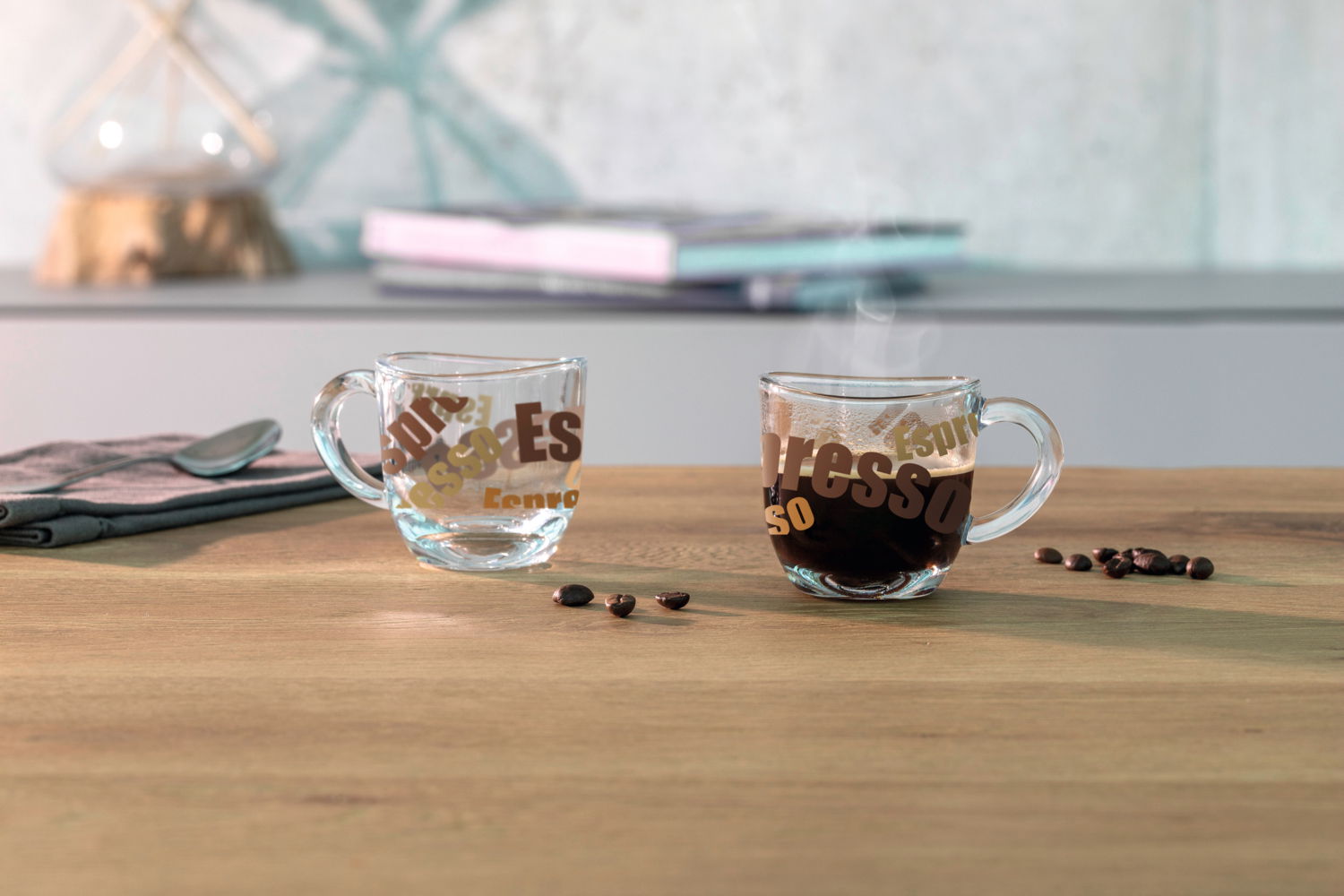 Zwei Espressotassen aus Glas mit buntem Schriftzug, eine gefüllt mit Espresso und dampfend, die andere leer, auf einem Holztisch mit Kaffeebohnen. Seitliche Perspektive.