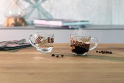 Zwei Espressotassen aus Glas mit buntem Schriftzug, eine gefüllt mit Espresso und dampfend, die andere leer, auf einem Holztisch mit Kaffeebohnen. Seitliche Perspektive.