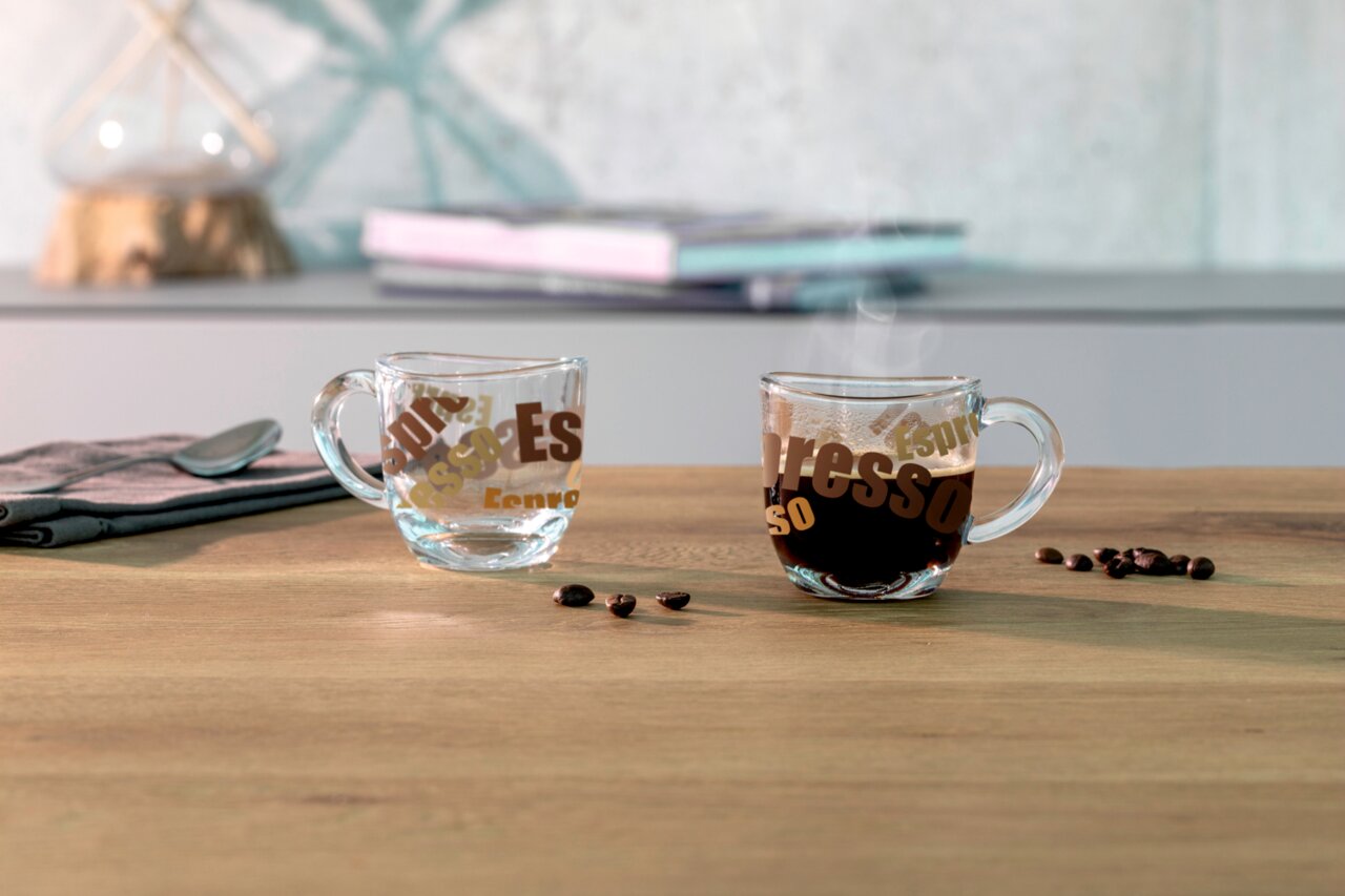 Zwei Espressotassen aus Glas mit buntem Schriftzug, eine gefüllt mit Espresso und dampfend, die andere leer, auf einem Holztisch mit Kaffeebohnen. Seitliche Perspektive.