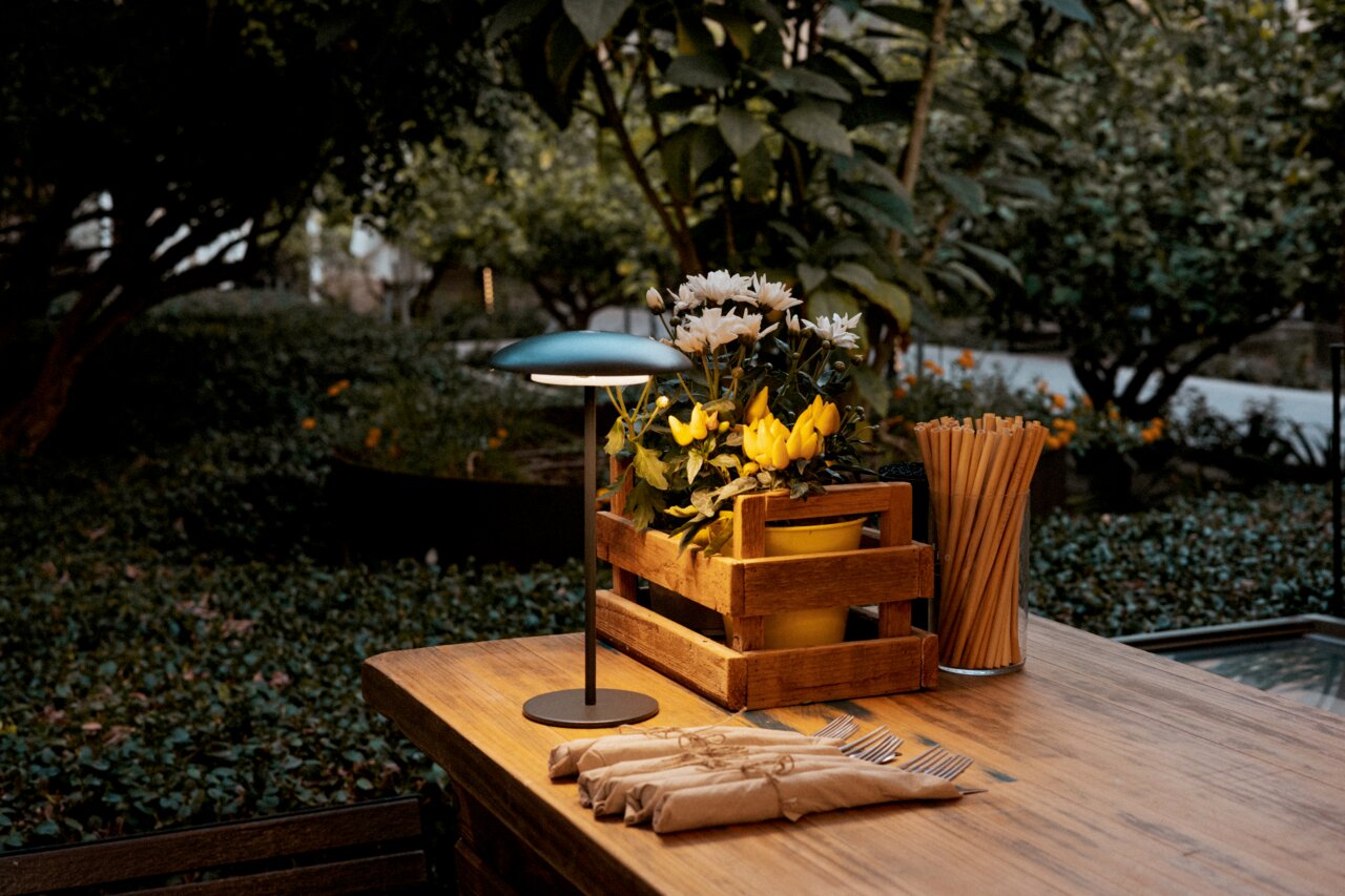 Een houten tafel in de tuin, schuin van voren gefotografeerd, met een kleine tafellamp, een houten kistje met gele en witte bloemen en een bakje met houten stokjes en ingepakt bestek.