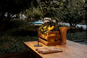 Een houten tafel in de tuin, schuin van voren gefotografeerd, met een kleine tafellamp, een houten kistje met gele en witte bloemen en een bakje met houten stokjes en ingepakt bestek.