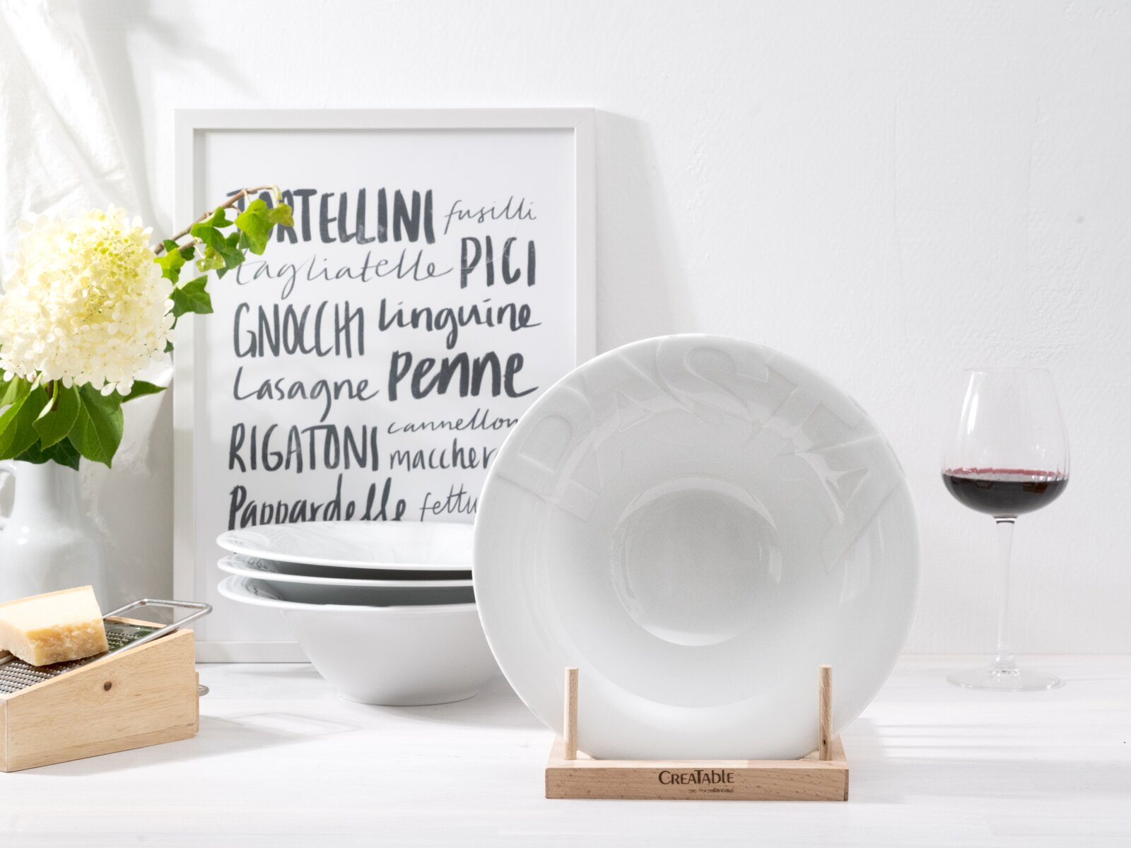 Creatable Pastateller 4-tlg GOURMET Weißer tiefer Porzellanteller/Schale auf Holzständer, frontal in leichter Seitenperspektive fotografiert; im Hintergrund gestapelte Schalen, Pasta-Poster und Rotweinglas.