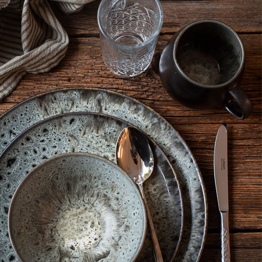 Rustikales Tischgedeck mit graublau gesprenkelten Keramiktellern und Schale, silbernem Löffel und Messer, Wasserglas und dunkler Tasse auf Holztisch, Draufsicht von oben
