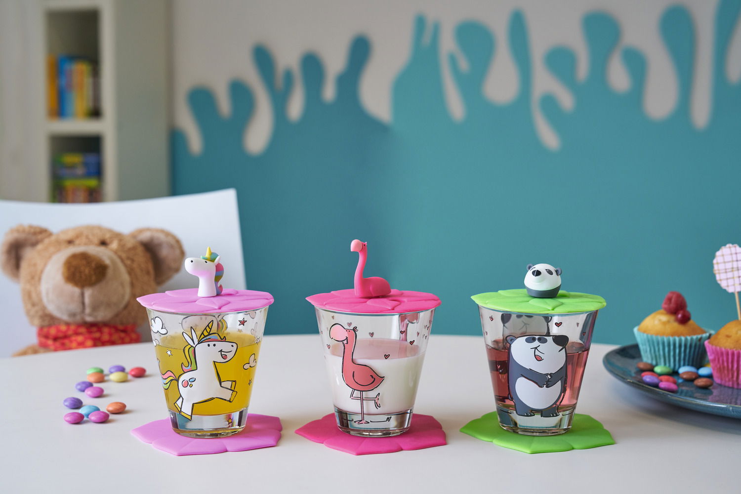 Drei bunte Kinder-Trinkgläser mit Tiermotiven und passenden Deckeln, fotografiert aus einer frontalen Perspektive. Ein Einhorn, ein Flamingo und ein Panda sind auf den Gläsern abgebildet, umgeben von bunten Süßigkeiten und einem Teddybär im Hintergrund.