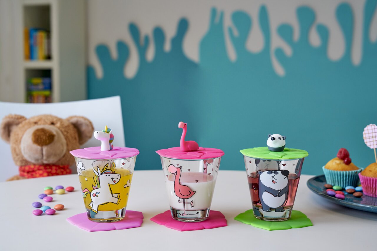Drei bunte Kinder-Trinkgläser mit Tiermotiven und passenden Deckeln, fotografiert aus einer frontalen Perspektive. Ein Einhorn, ein Flamingo und ein Panda sind auf den Gläsern abgebildet, umgeben von bunten Süßigkeiten und einem Teddybär im Hintergrund.