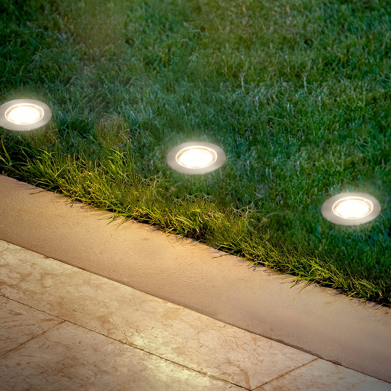 Bodenleuchten aus Edelstahl, eingebettet in Rasen neben einer Terrasse, aus der Vogelperspektive fotografiert.