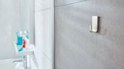 Nahaufnahme eines an der Badezimmerwand befestigten tesa Powerstrips Waterproof Hakens, mit unscharfem Hintergrund eines Waschbeckens und Zahnbürstenhalters.