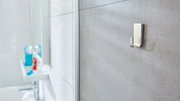 Nahaufnahme eines an der Badezimmerwand befestigten tesa Powerstrips Waterproof Hakens, mit unscharfem Hintergrund eines Waschbeckens und Zahnbürstenhalters.
