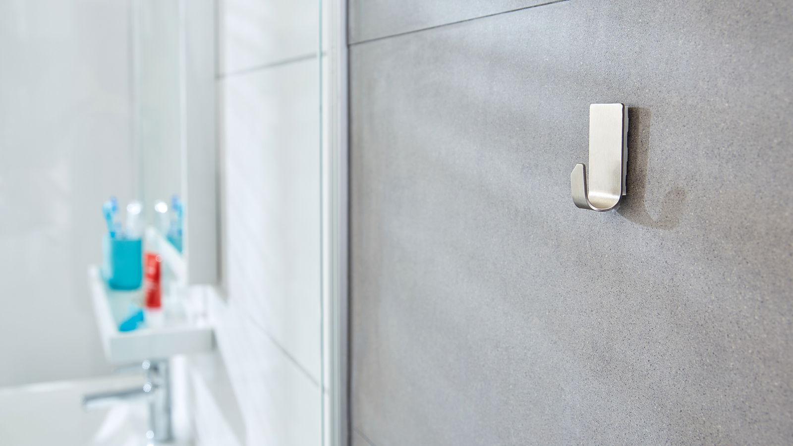 Nahaufnahme eines an der Badezimmerwand befestigten tesa Powerstrips Waterproof Hakens, mit unscharfem Hintergrund eines Waschbeckens und Zahnbürstenhalters.