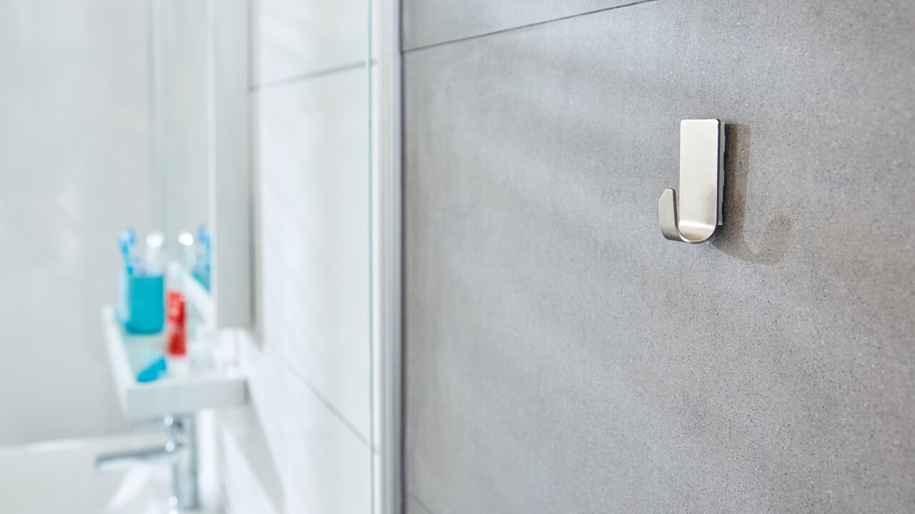 Nahaufnahme eines an der Badezimmerwand befestigten tesa Powerstrips Waterproof Hakens, mit unscharfem Hintergrund eines Waschbeckens und Zahnbürstenhalters.