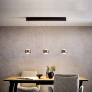 Moderne hanglamp met drie lampen, voorzien van drie ronde metalen lampenkappen, bevestigd aan een zwarte plafondrail boven een eettafel; vooraanzicht, licht van onderaf