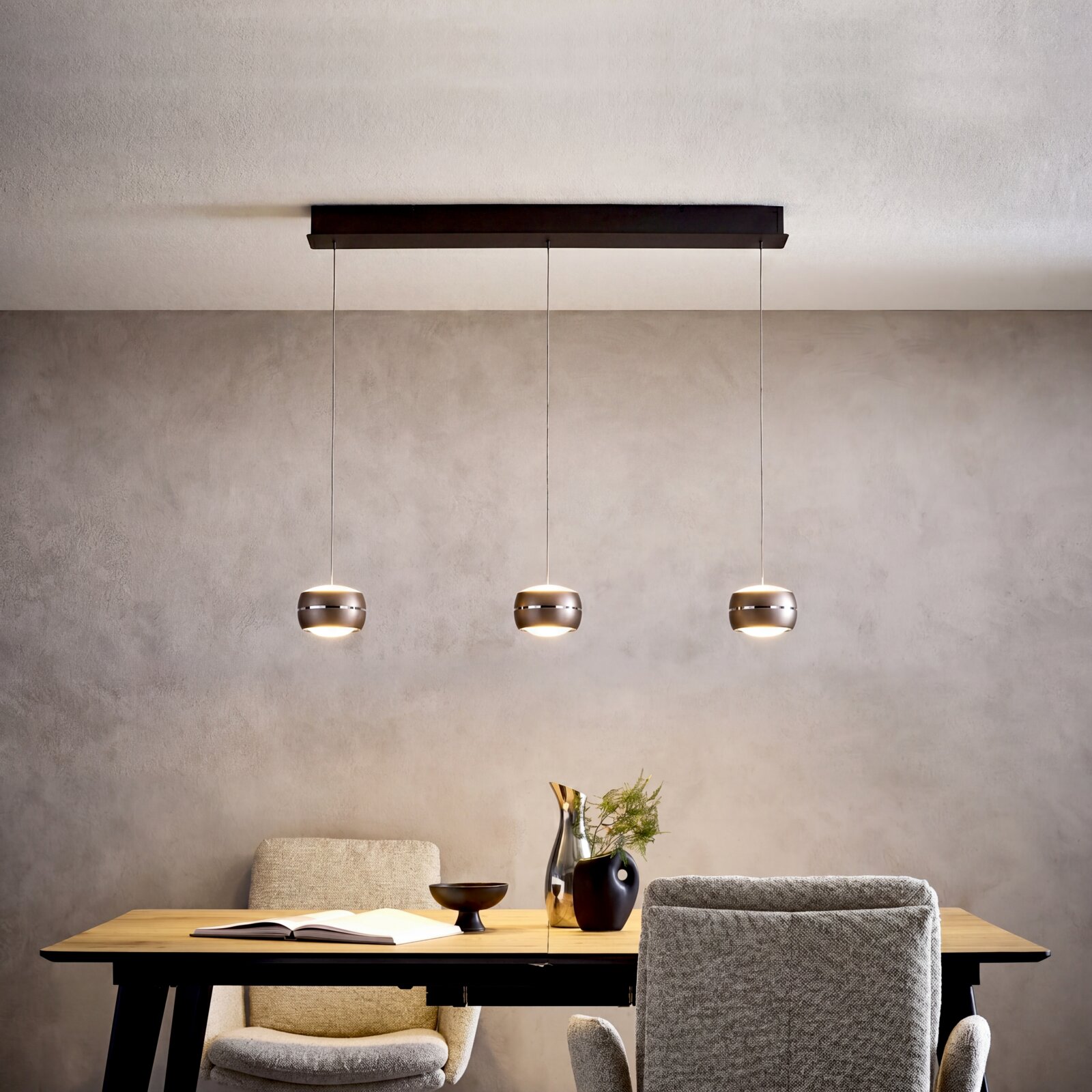Moderne hanglamp met drie lampen, voorzien van drie ronde metalen lampenkappen, bevestigd aan een zwarte plafondrail boven een eettafel; vooraanzicht, licht van onderaf