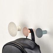 Drei Wandhaken in verschiedenen Farben, von links nach rechts: beige, rosa und grau, an einer Wand montiert. Ein schwarzer Rucksack hängt am mittleren Haken. Perspektive von vorne.