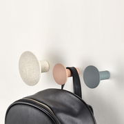 Drei Wandhaken in verschiedenen Farben, von links nach rechts: beige, rosa und grau, an einer Wand montiert. Ein schwarzer Rucksack hängt am mittleren Haken. Perspektive von vorne.